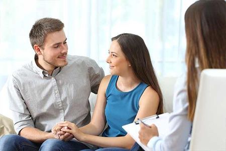 A man and a woman are sitting on a couch holding hands and talking to a woman.