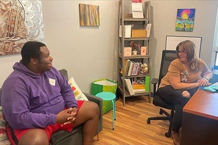 A man in a purple hoodie is sitting on a couch talking to a woman in an office.