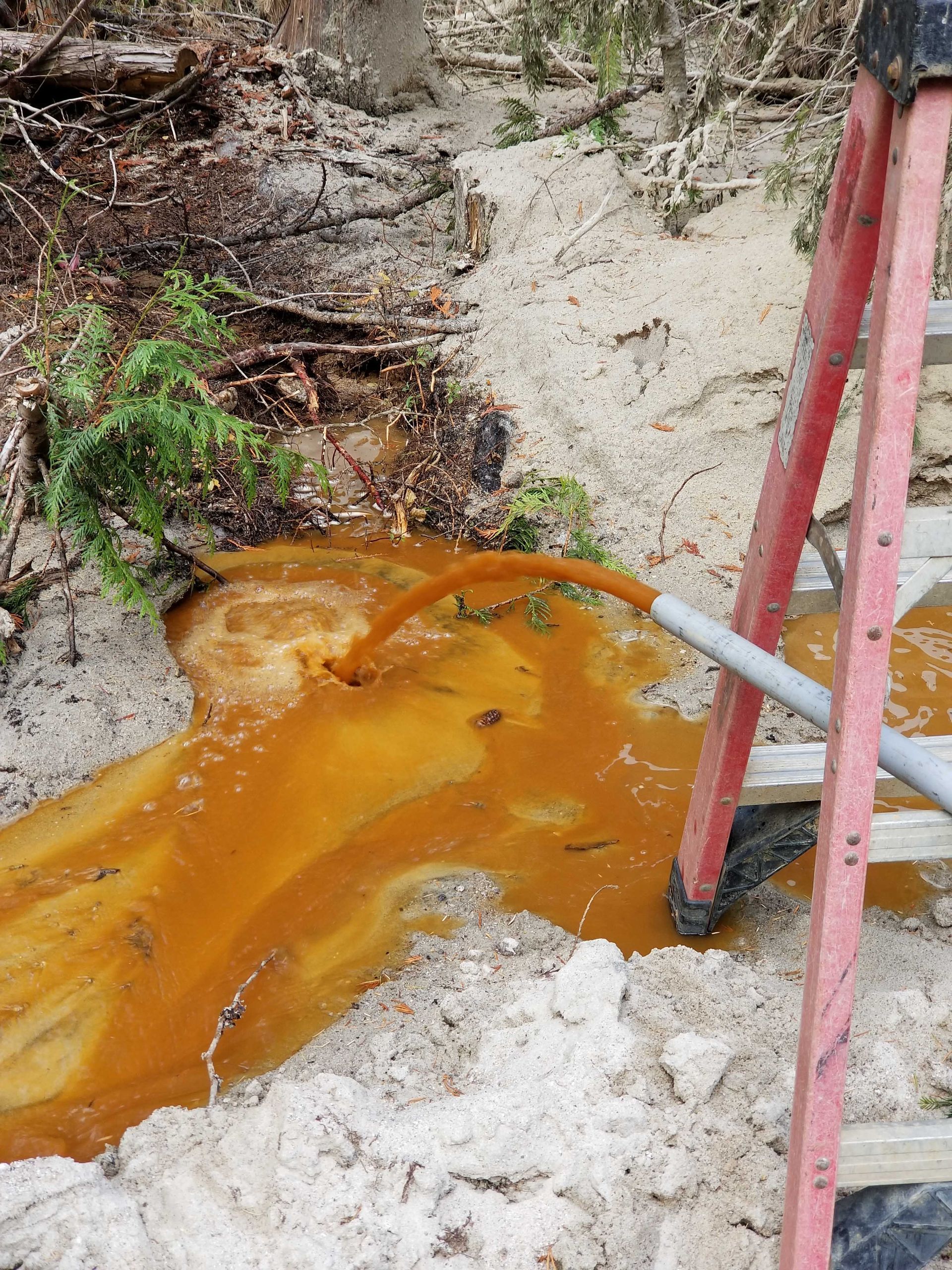 Orange-colored water pours from a pipe onto sandy ground near a ladder and greenery.