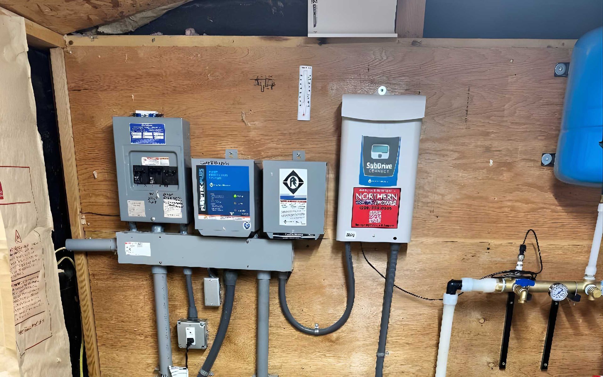 Electrical control panel with multiple boxes and wiring on a wood wall; a blue pressure tank is to the right.