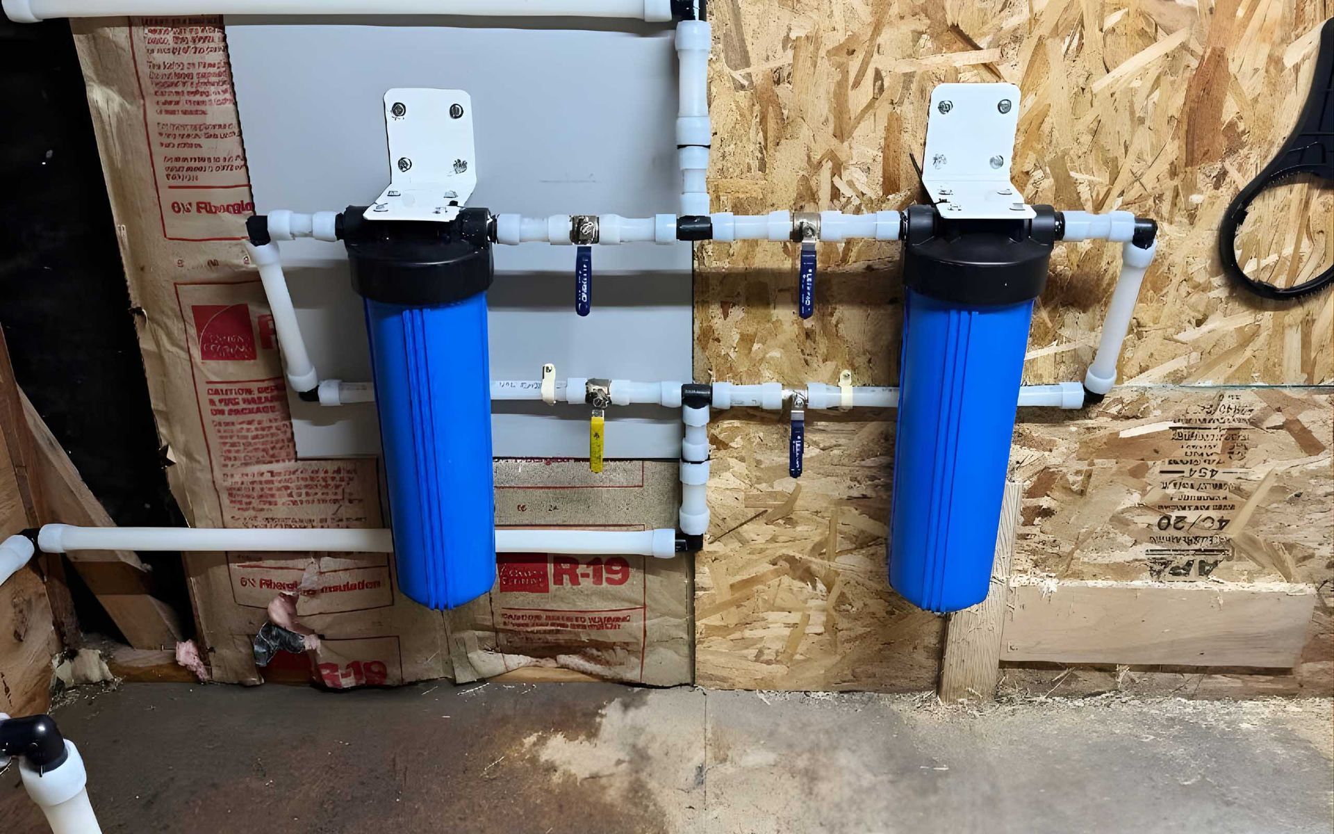 Two blue water filter housings with white PVC pipes and valves on a wood wall.