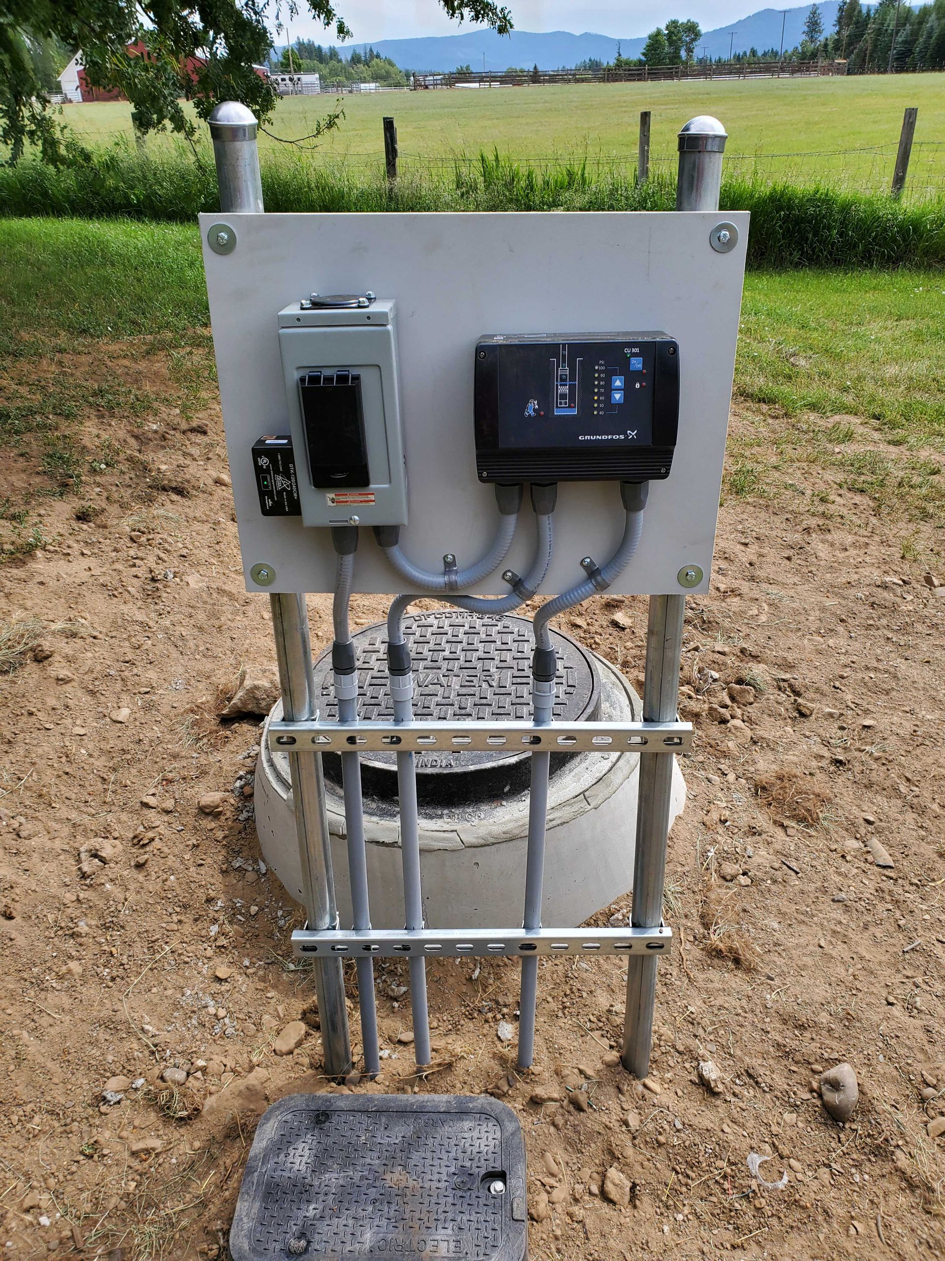 Electrical box mounted on supports above a circular concrete well, field in background.