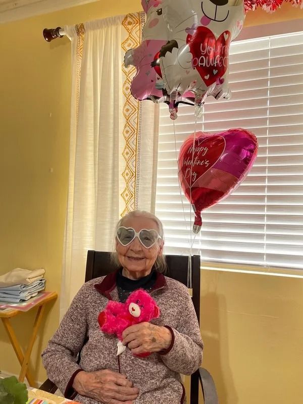 Elderly woman wearing heart-shaped glasses, holding a pink plush bear, with Valentine's Day balloons in the background. She is smiling in a warmly lit room.