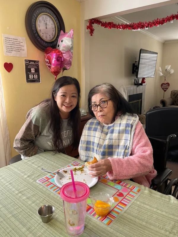 Woman and elderly woman in pink robe seated at a table, one eating, with decorations including balloons and hearts.