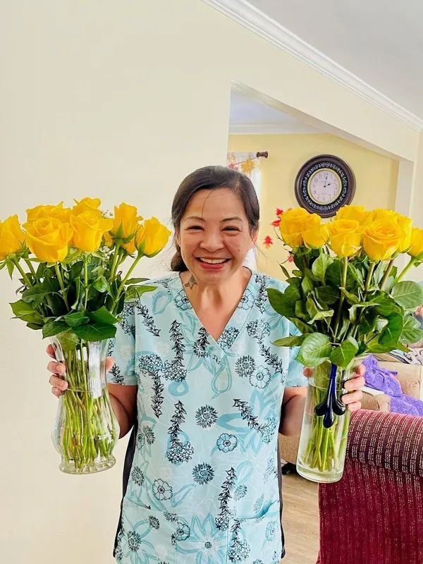 Woman holding two bouquets of yellow roses, smiling. She's wearing a light blue patterned top inside a home.