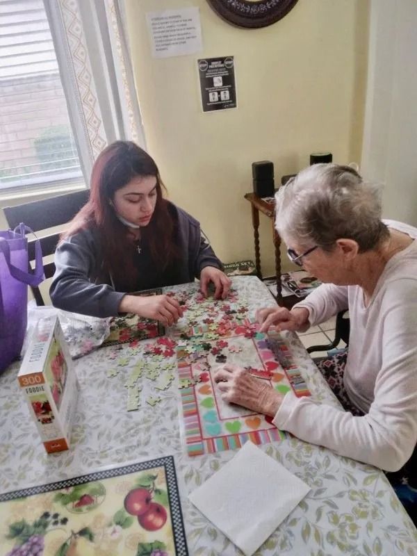 A young woman and an elderly woman with glasses sit at a table, working on a jigsaw puzzle together.