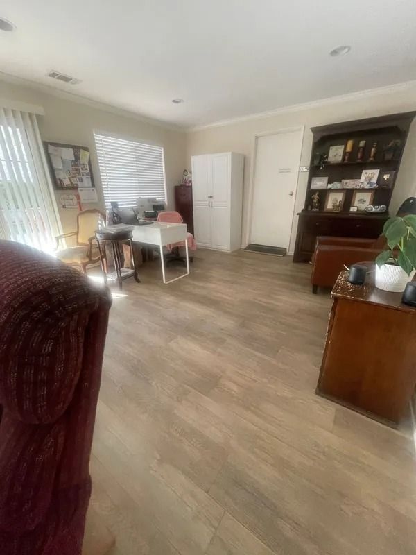 An office with light wood flooring, a white cabinet, a dark wood cabinet, a desk by a window, and a burgundy armchair.