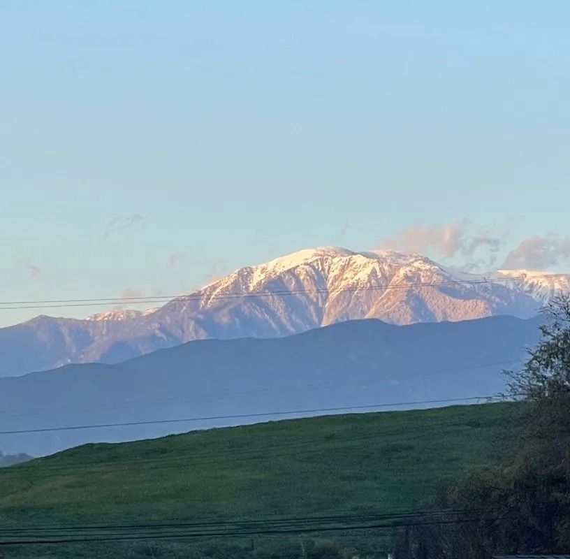 Snow-capped mountains at sunrise, with a green field in the foreground under a light blue sky.
