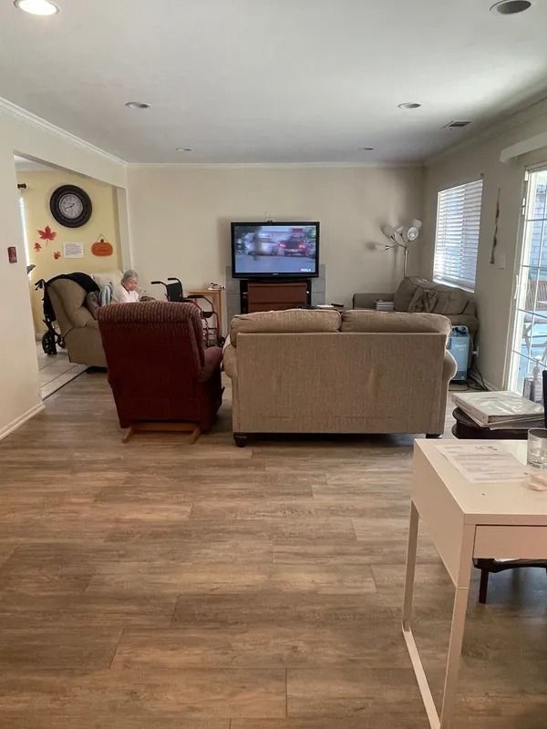 Living room with a TV, two couches, and a recliner. A person is visible in the adjacent room. The floors are a light wood color.