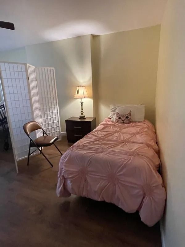 A small bedroom with a pink-covered bed, nightstand, and folding screen. A lamp is on the nightstand, and a chair sits nearby.