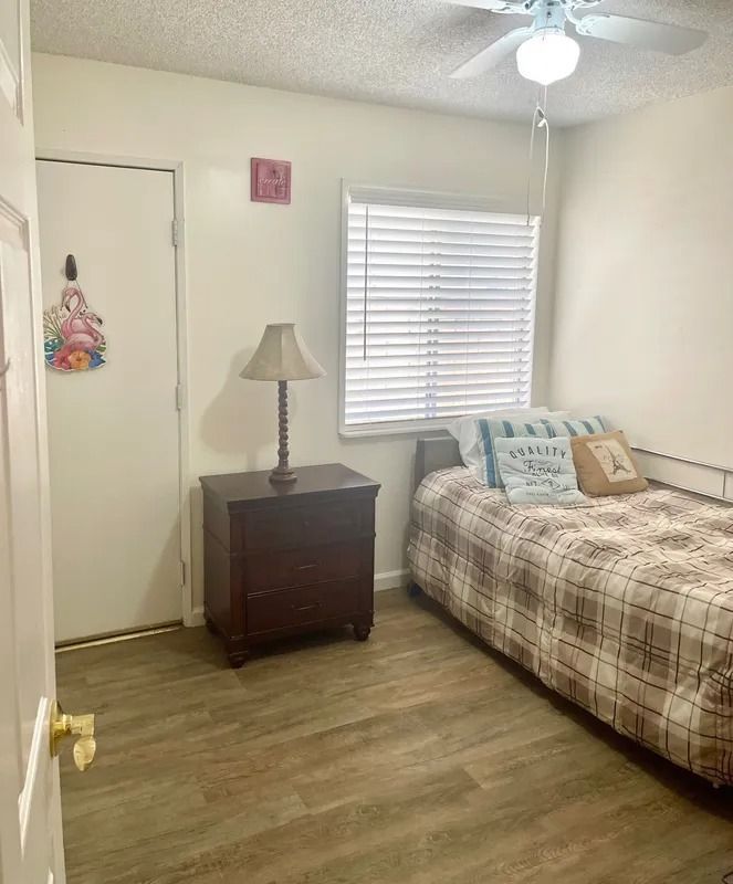 Bedroom with a bed, nightstand, door, and window with blinds. Light-colored walls and wood-look flooring.