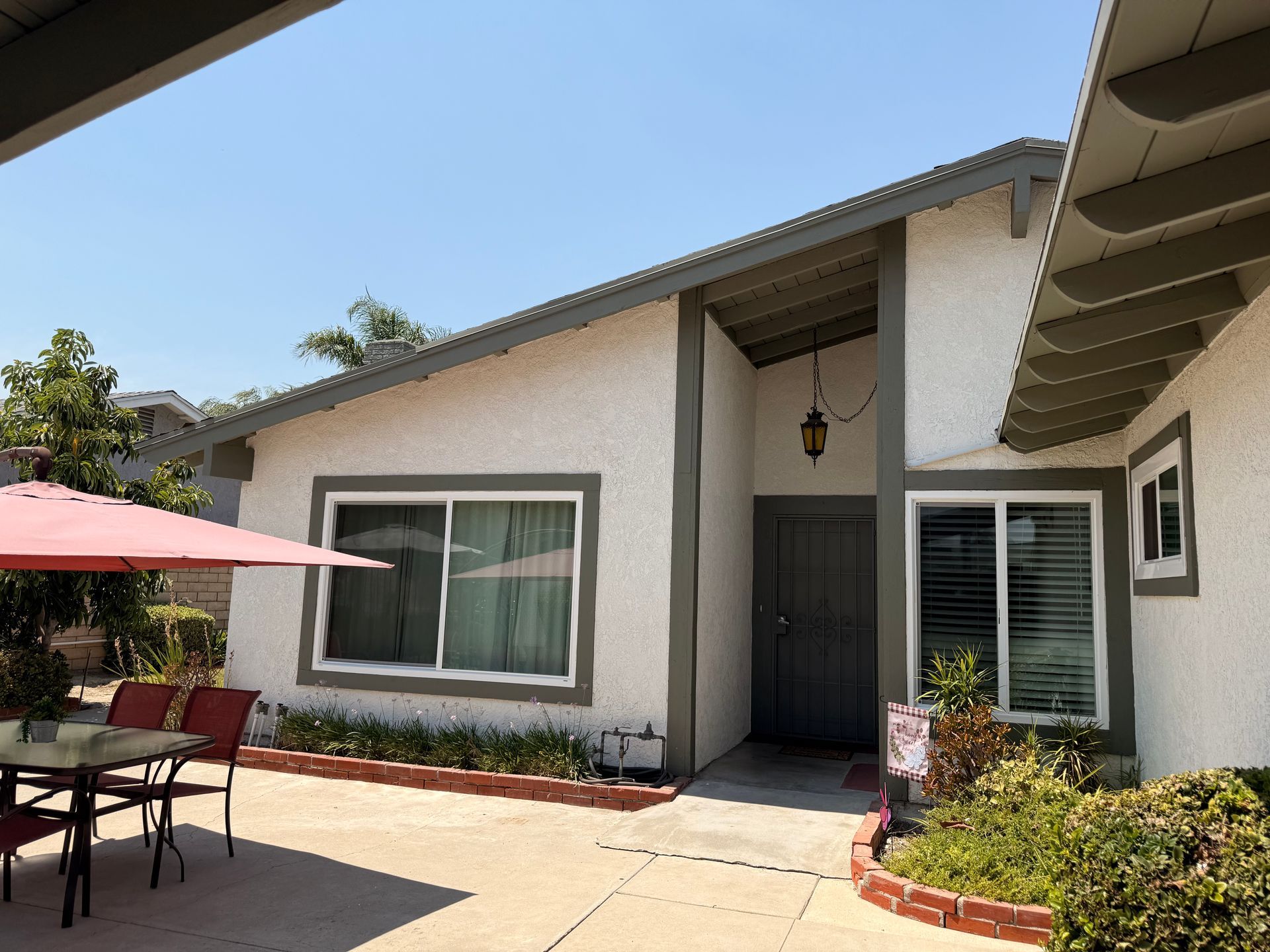 Exterior view of a single-story house with white stucco walls, grey trim, and a small patio with a table and umbrella.