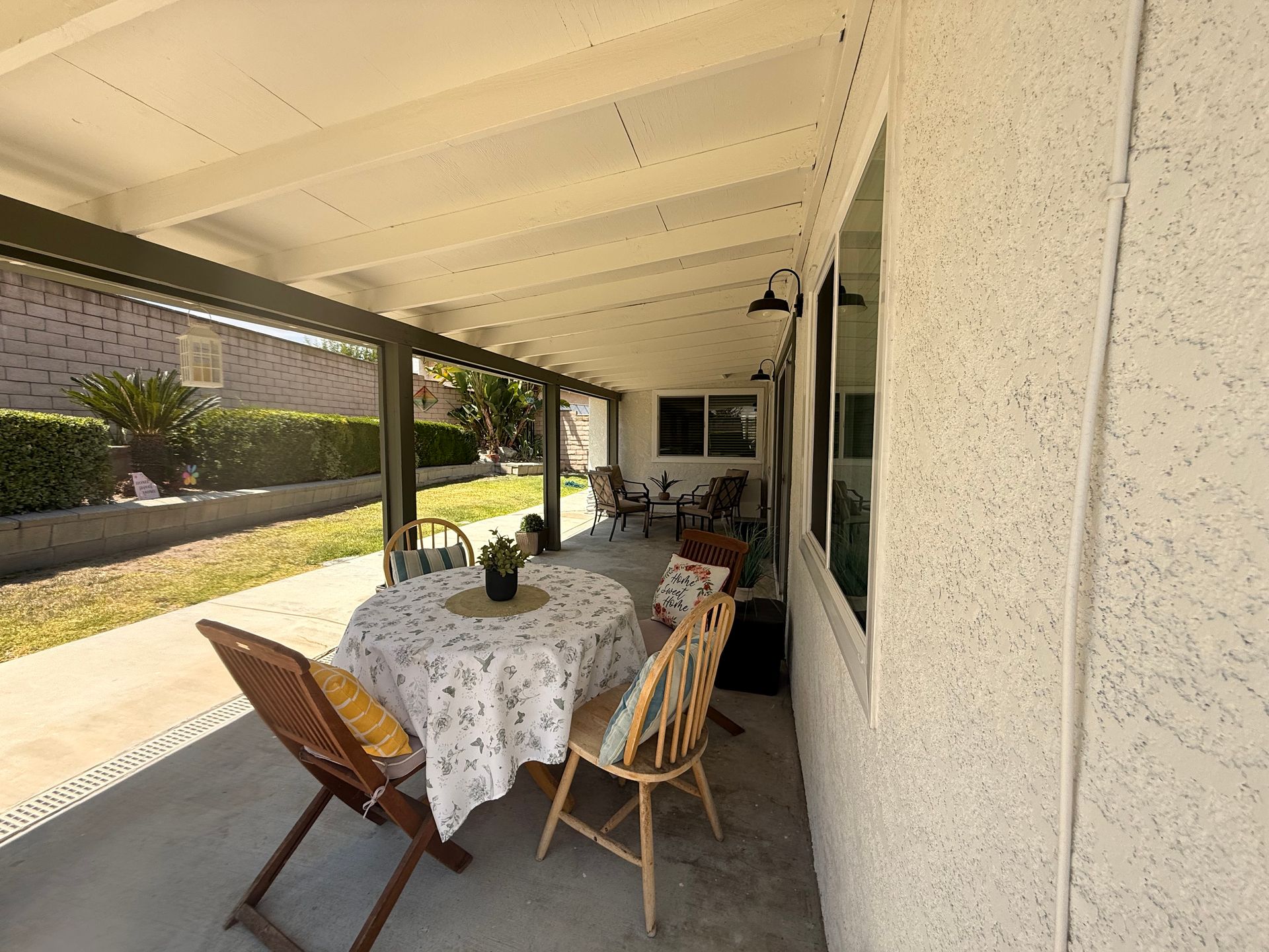 Covered patio with a round table set for a meal. Wooden chairs surround the table, with a floral tablecloth.