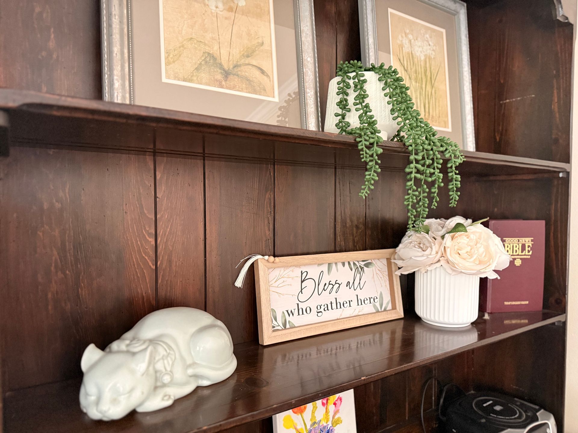Wooden bookshelf with framed art, a white cat figurine, a sign that says 