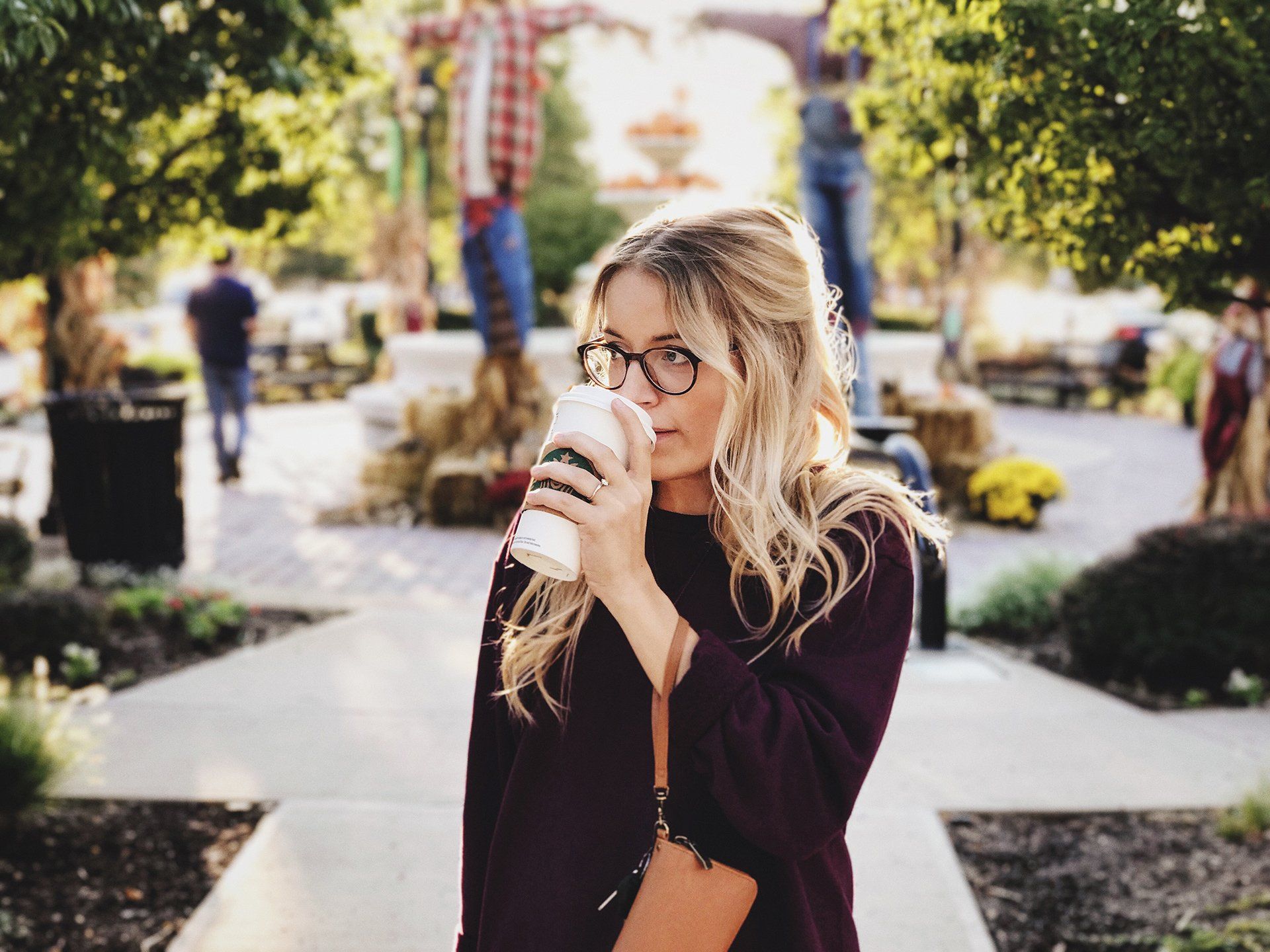 Woman with glasses drinks from a coffee cup outdoors; fall setting with scarecrows and foliage.
