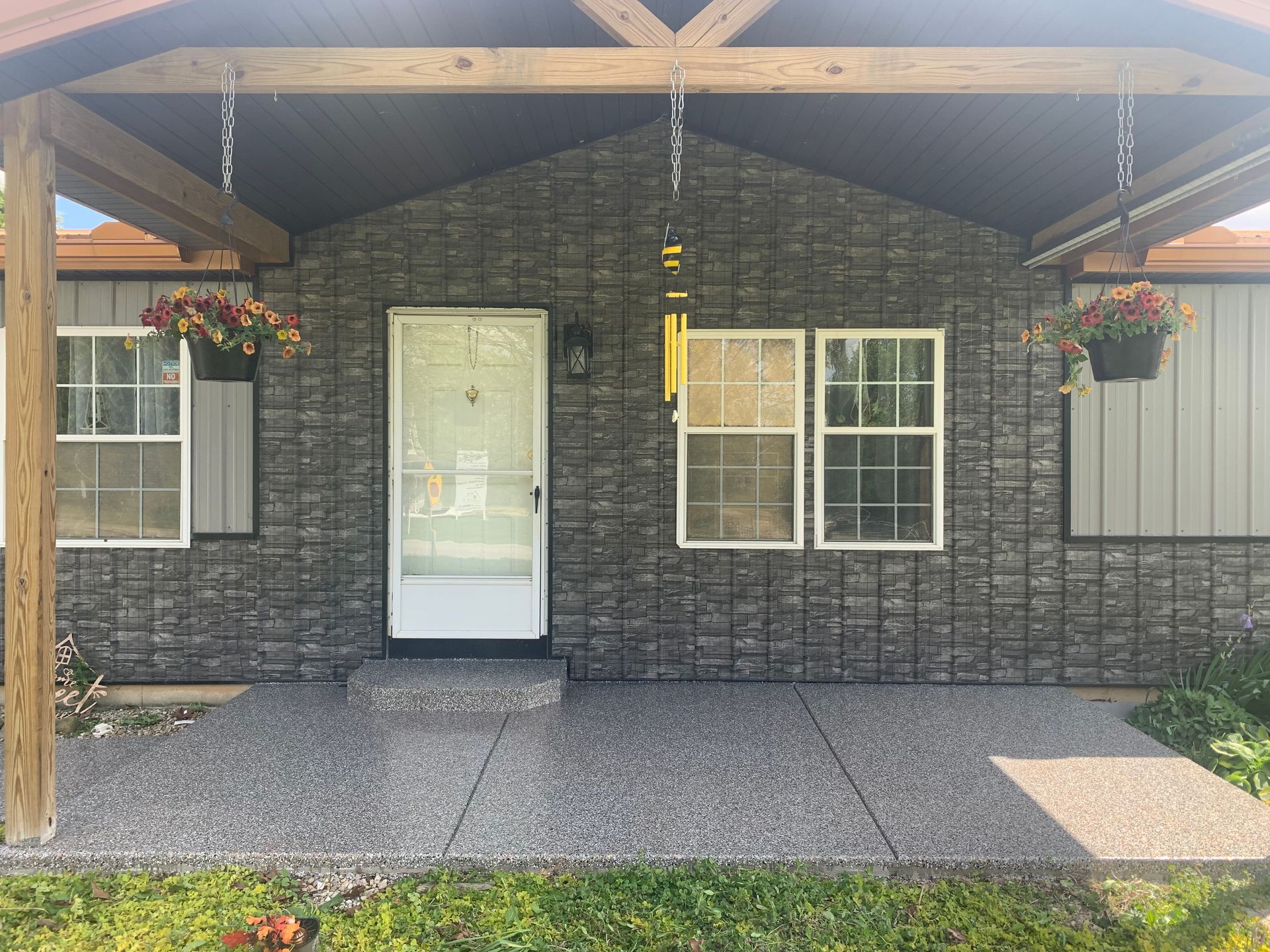 Front porch with gray stone facade, white door and window, wooden beams, hanging flower baskets, and gravel walkway.