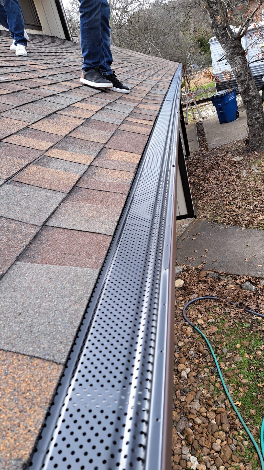 Person on a rooftop by a gutter with a metal mesh cover. Shingled roof, brown and grey.