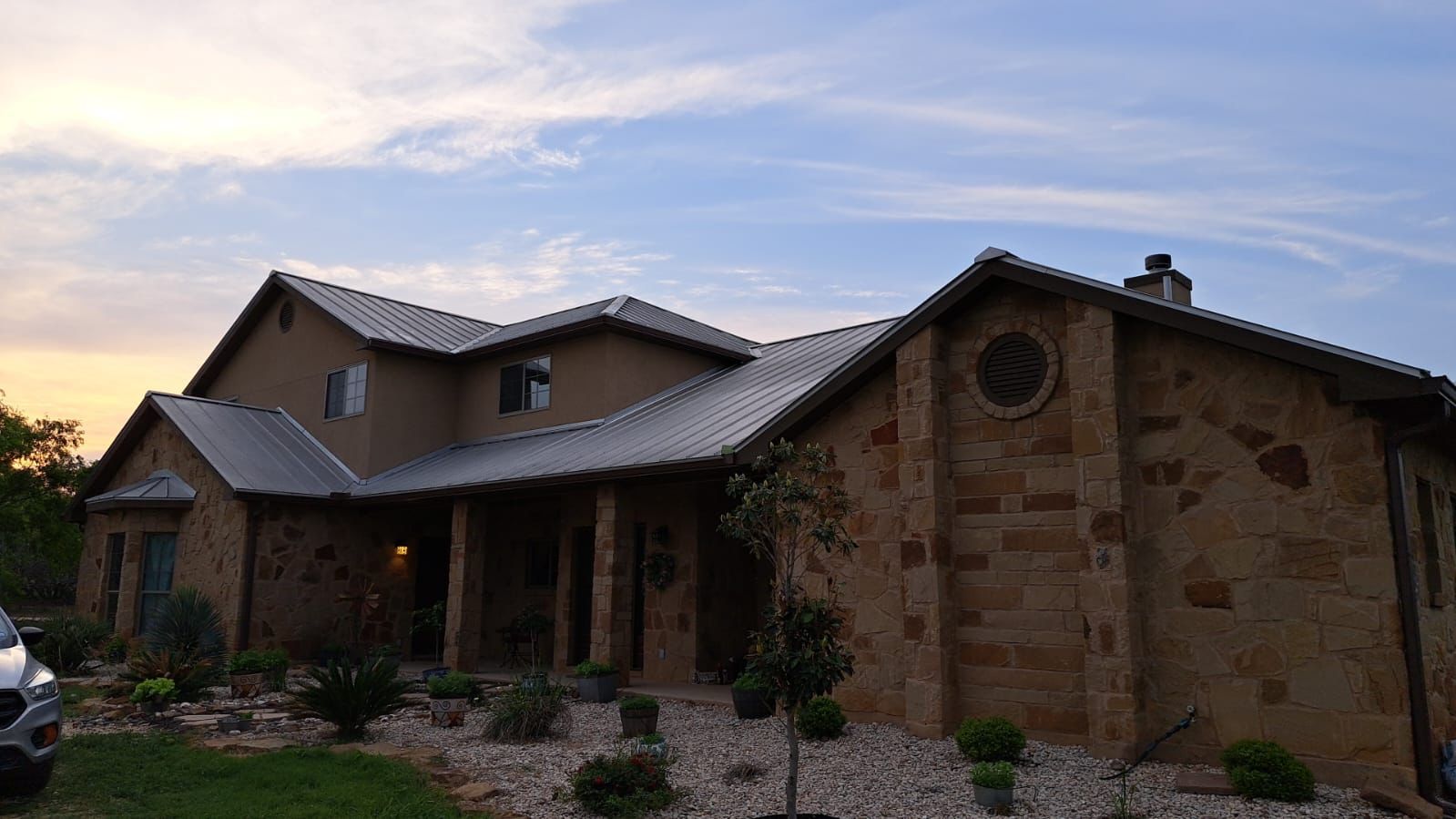 Stone house with metal roof, porch, and a circular window, under a dusk sky.