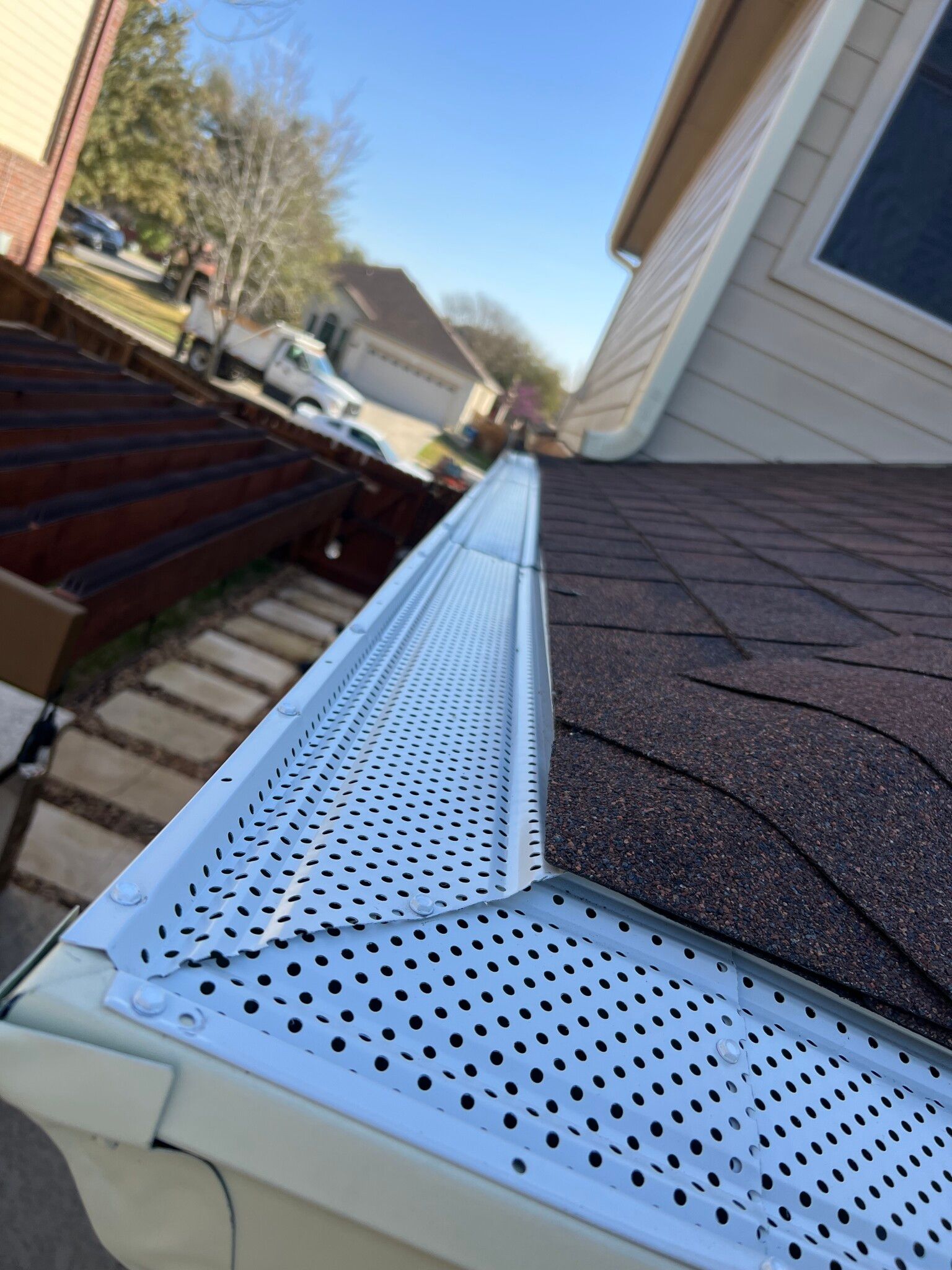 White gutter guard installed on a brown shingled roof, angled view under a blue sky.