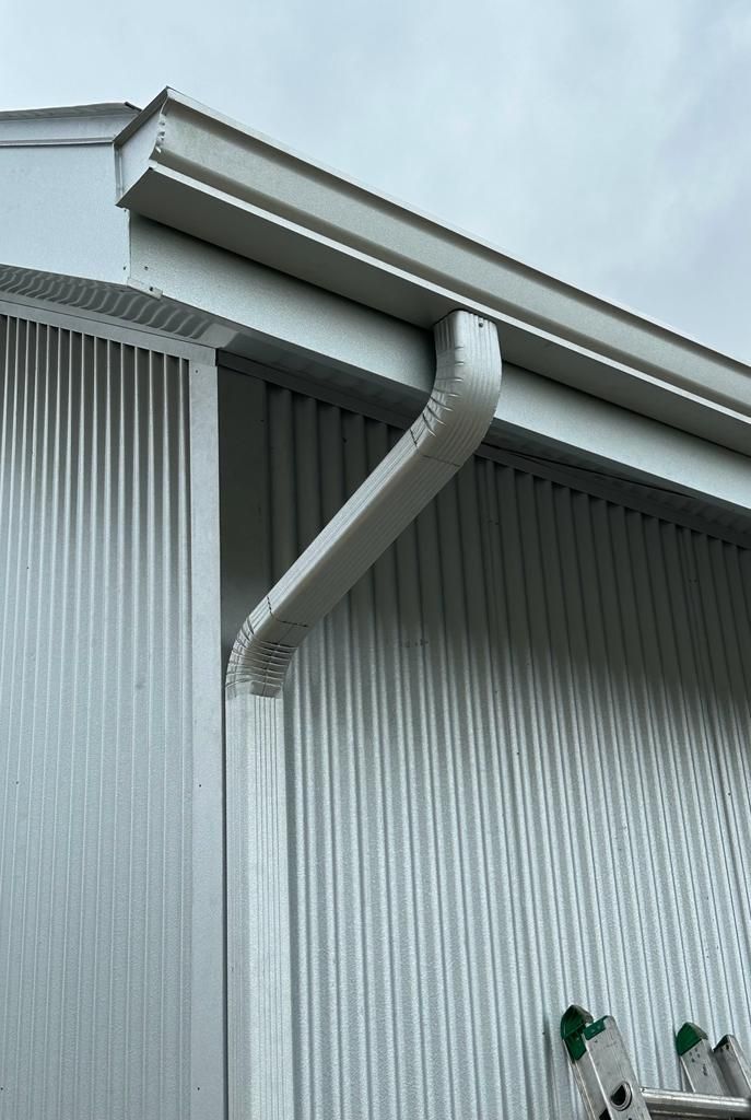 Metal building with a gutter and downspout on the side, against a cloudy sky.