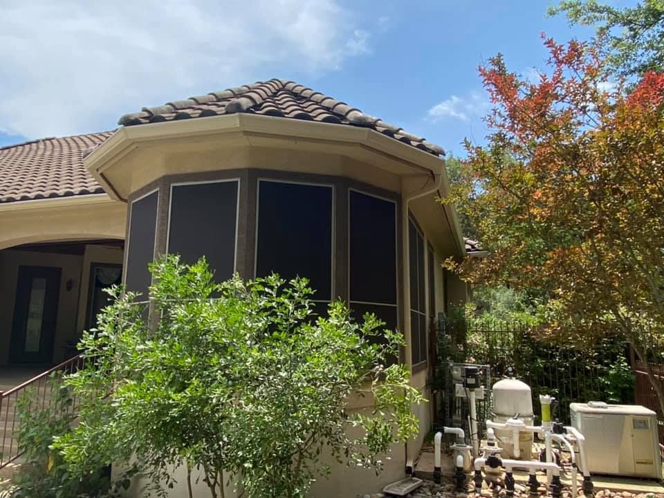 Screened-in patio with dark screens, beige structure, and brown roof. Green plants and pool equipment are visible.