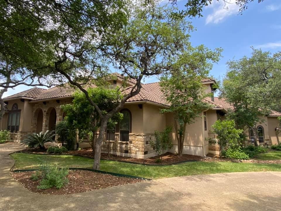 Beige stucco house with brown tile roof, stone accents, and lush landscaping under a partly cloudy sky.