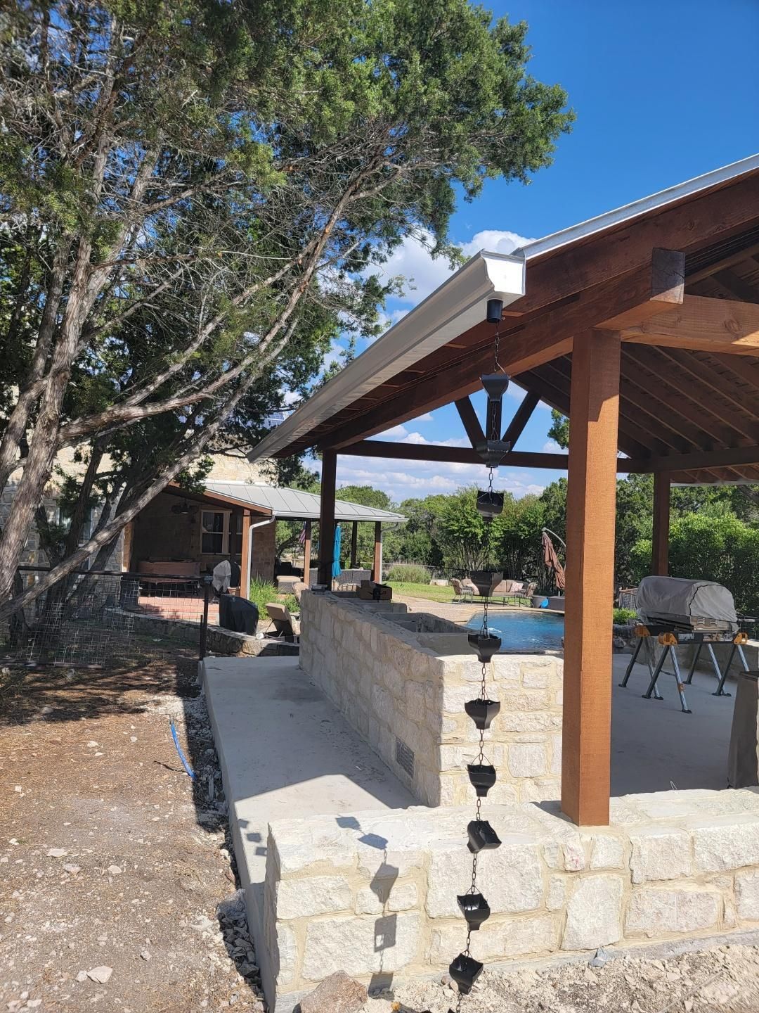 Outdoor kitchen with a stone wall and wooden pergola near a pool under a blue sky.