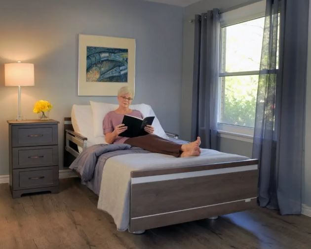 Woman reading in adjustable bed, gray bedroom with window, nightstand, artwork.