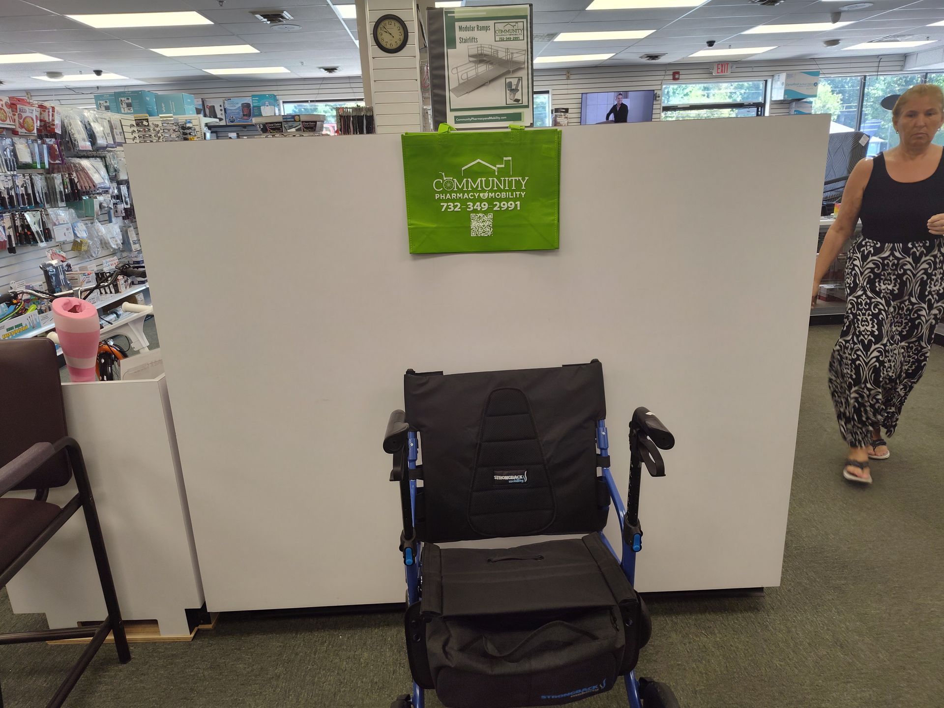 A blue and black rolling walker is in a store aisle in front of a white partition. A woman walks by.