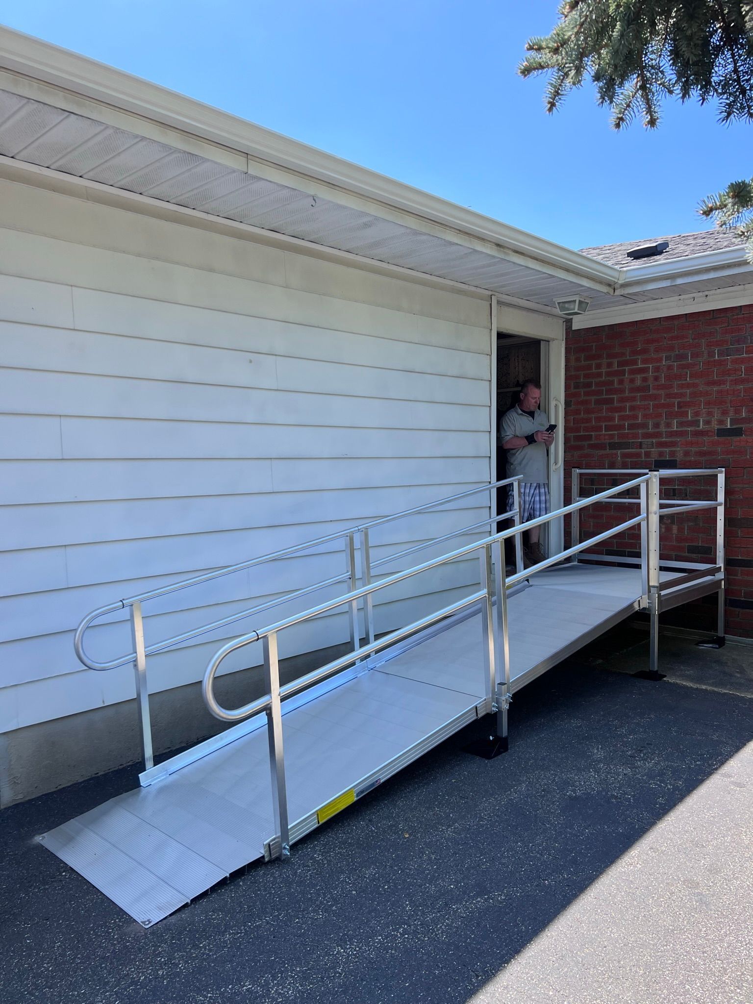 An aluminum ramp with handrails provides wheelchair access to a building; a person stands in the doorway.