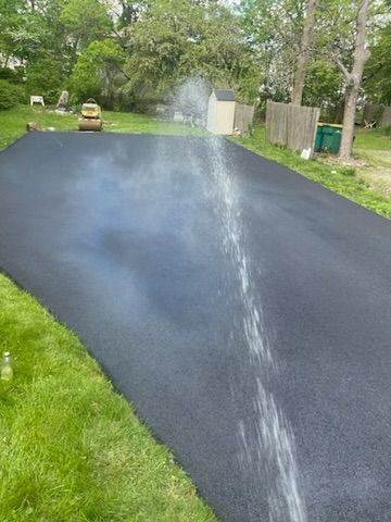 Newly paved black asphalt driveway being sprayed with water, surrounded by green grass and a suburban setting.