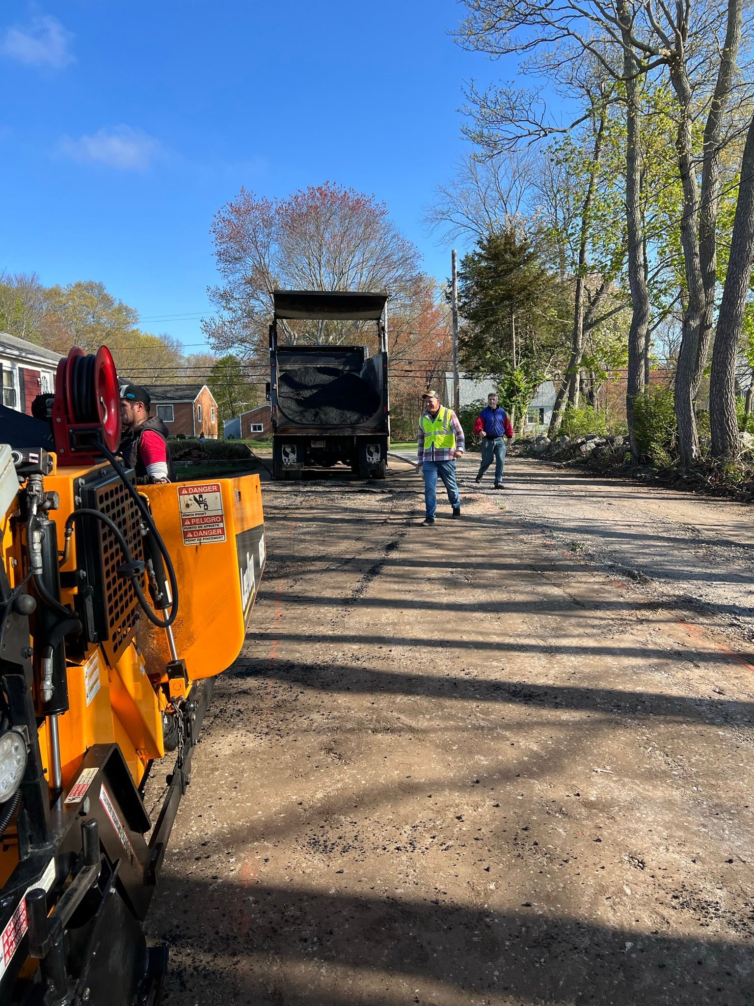 Paving crew at work; asphalt truck, machinery, and workers on a paved road under blue sky.