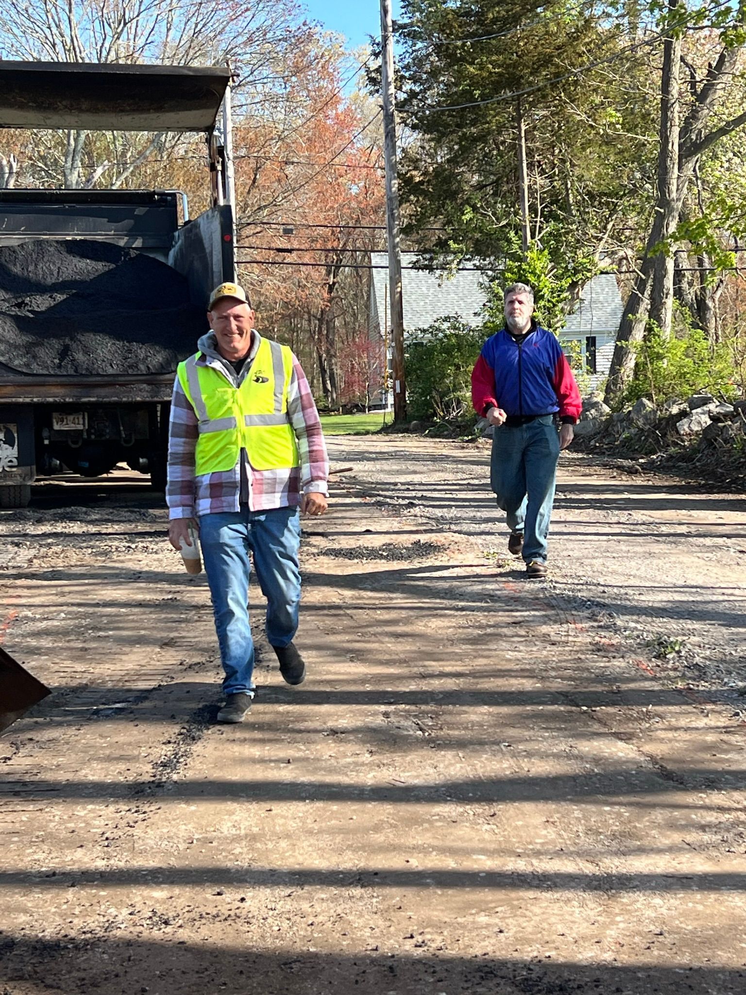 Two men walk on a gravel road near a truck. One wears a yellow vest, the other a blue shirt. Autumn setting.