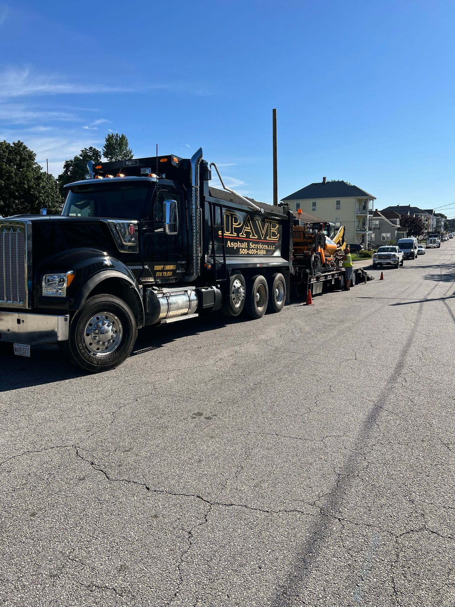 Black semi-truck with trailer carrying equipment parked on a street. Houses and cars are in the background on a sunny day.