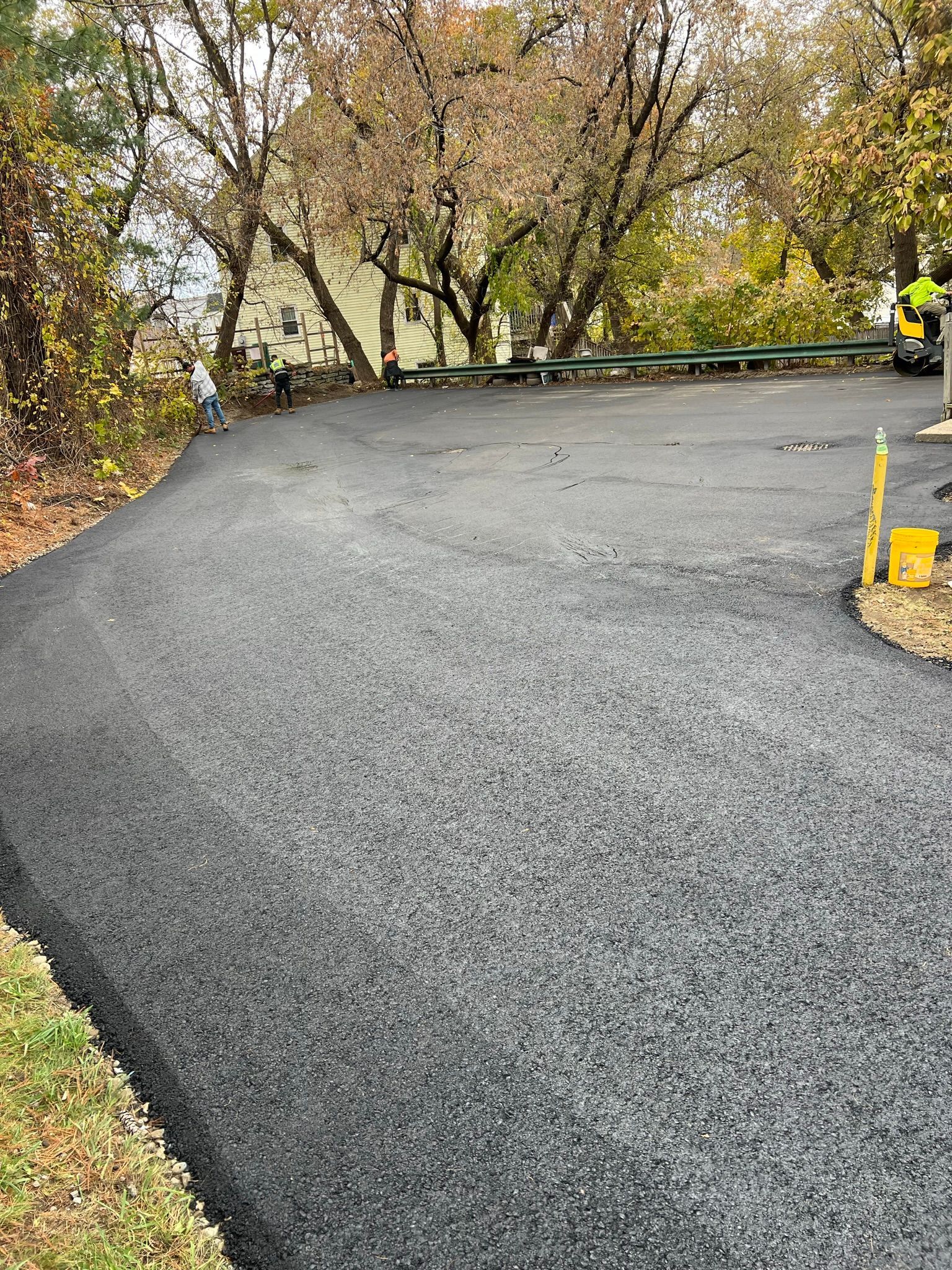 Newly paved asphalt driveway curving toward trees and a house on a fall day.