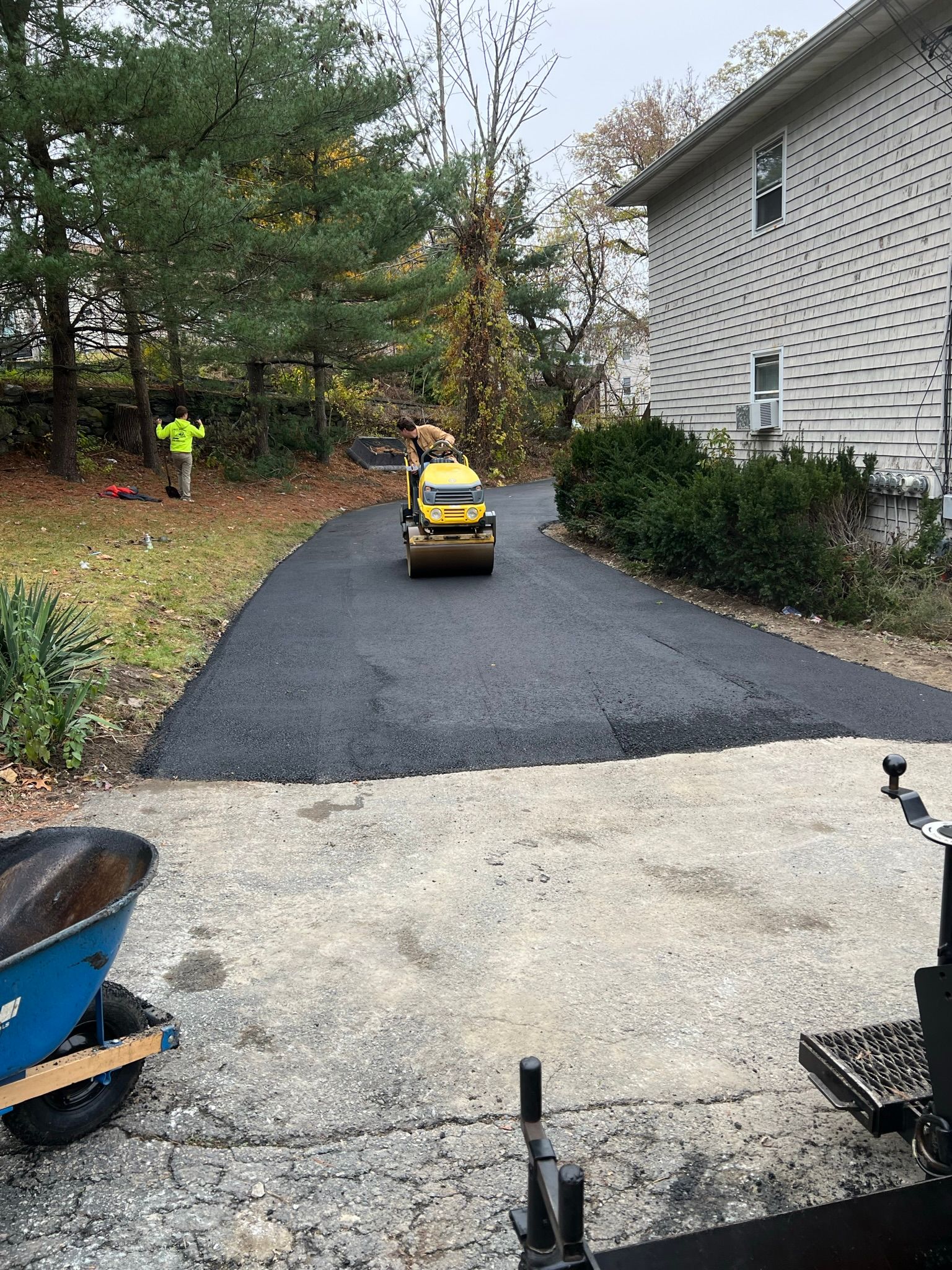Asphalt driveway being paved with roller machine; worker in safety vest nearby.
