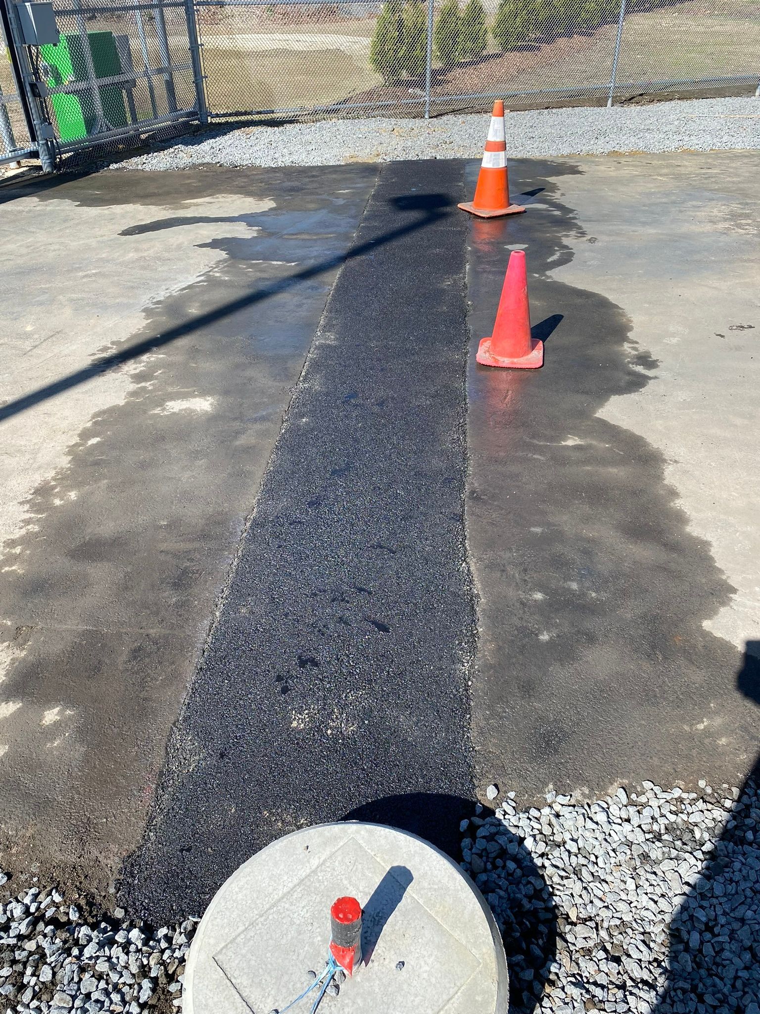 Strip of asphalt repair on concrete, marked by cones. Gravel surrounds the repair on wet pavement.