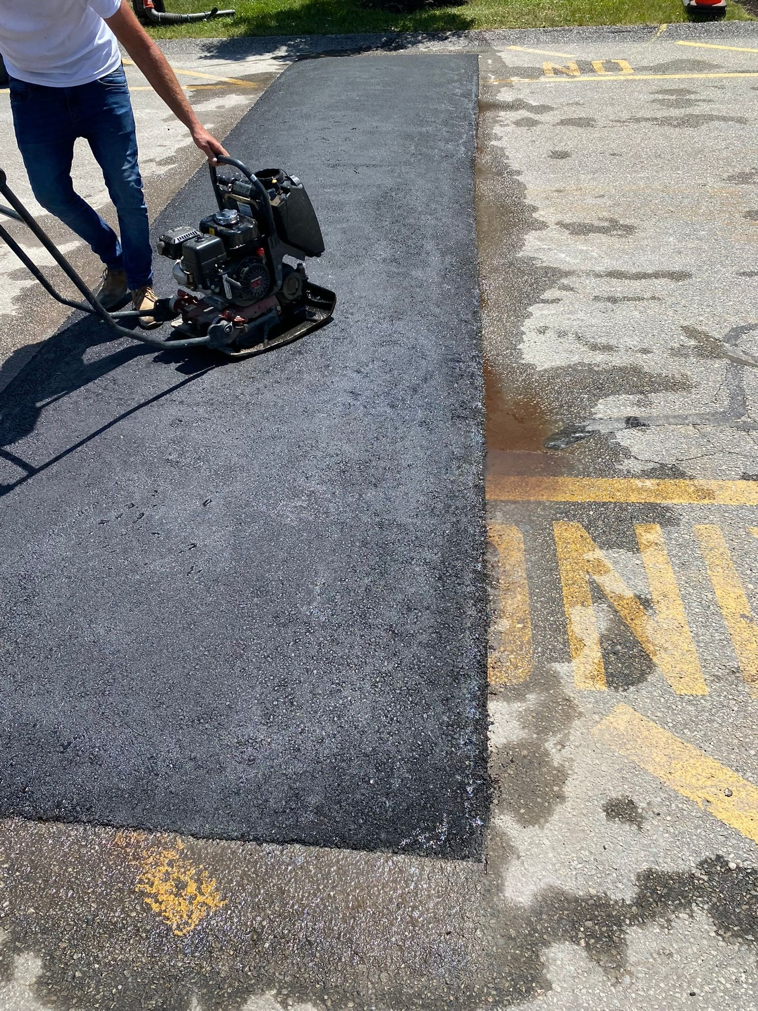 Man using a plate compactor on fresh asphalt in a parking lot, next to painted yellow lines.