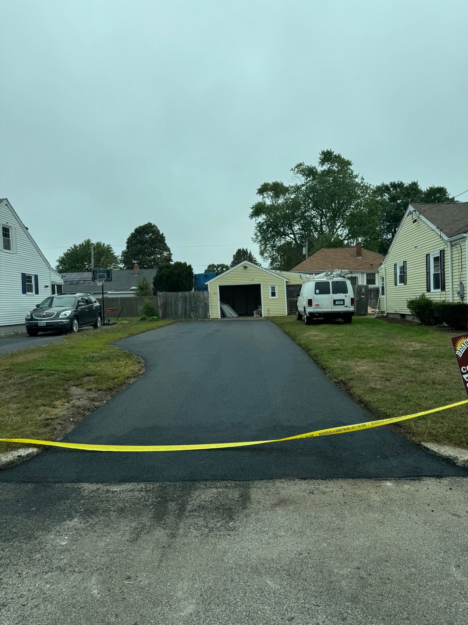 Black asphalt driveway with yellow caution tape and two houses on either side.