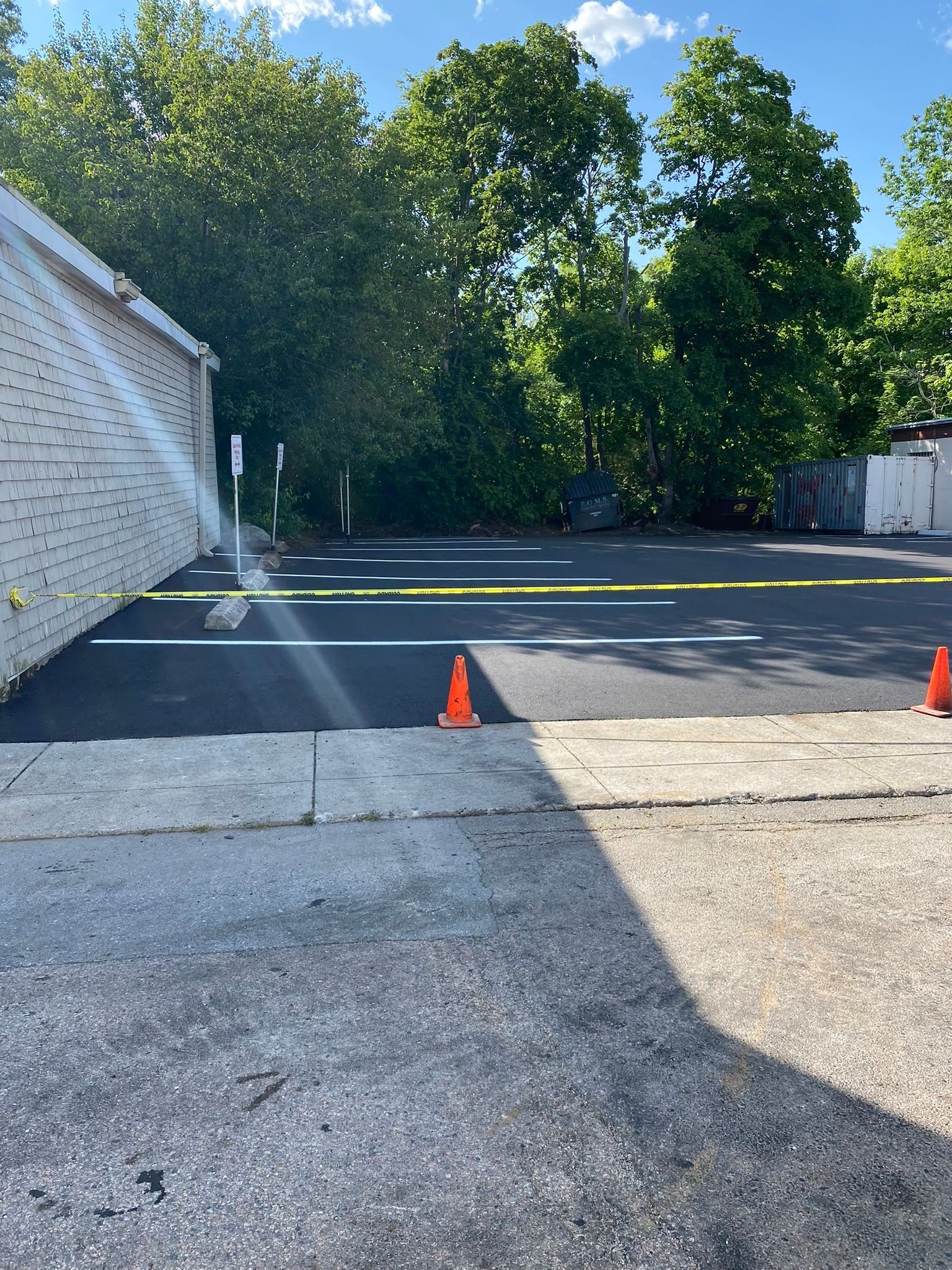 Newly paved parking lot, with white lines, yellow tape, orange cones. Trees in background.