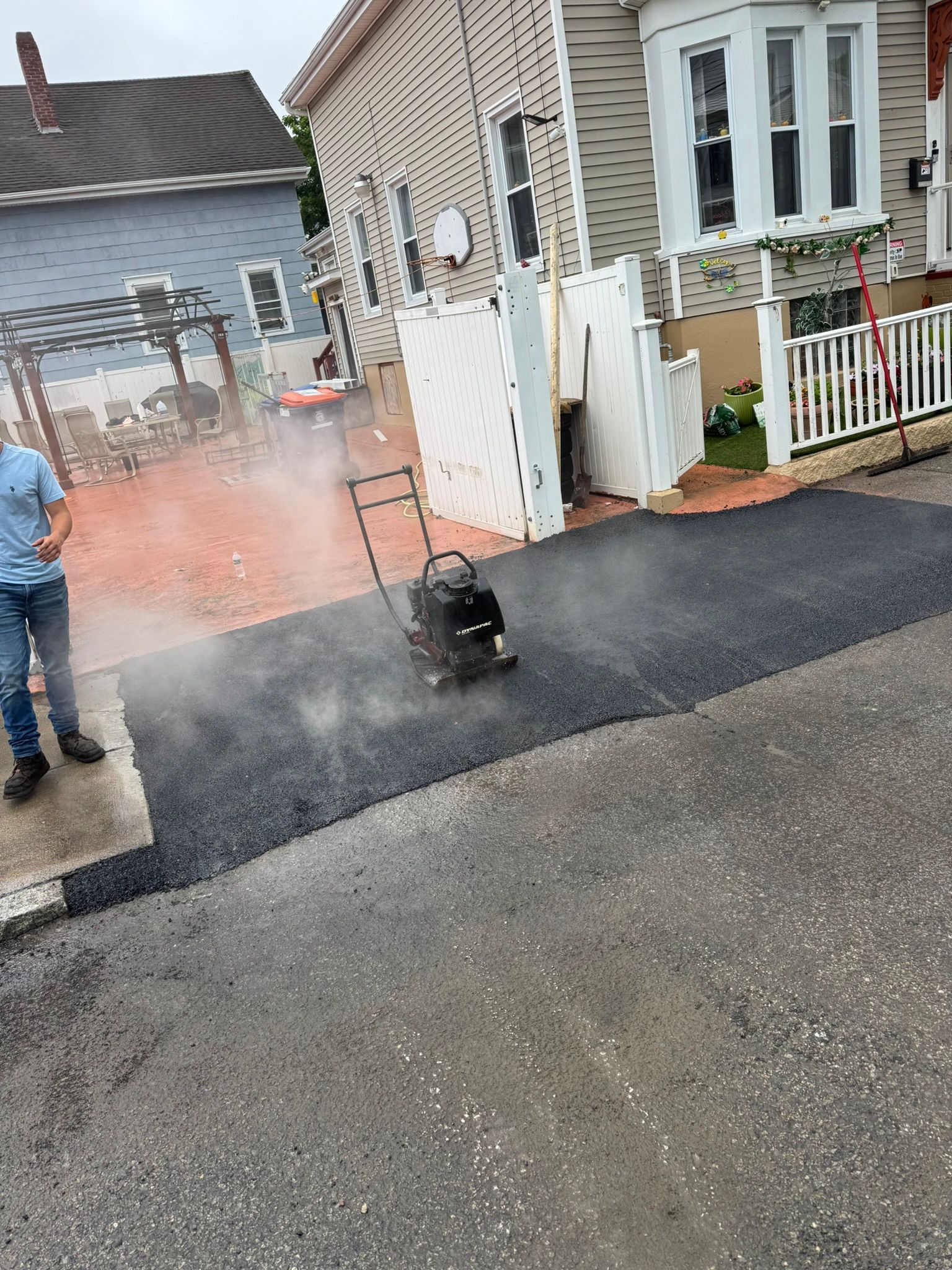 Asphalt paving in progress; a worker operates a compactor. Steam rises from fresh asphalt near buildings.