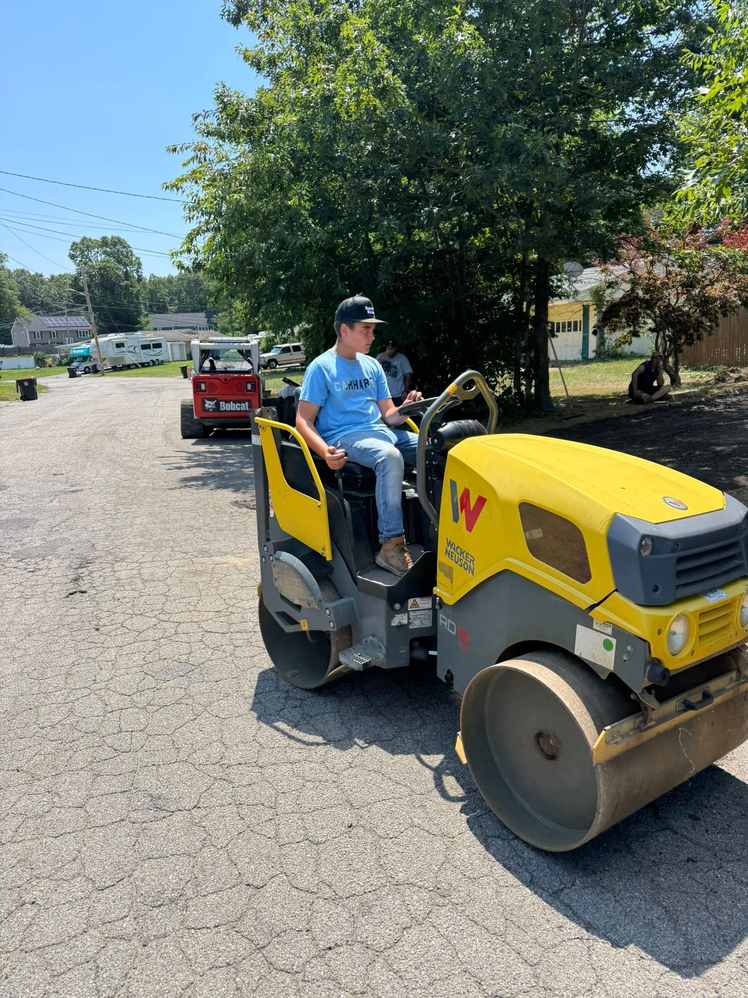 Man operating a yellow and black Wacker Neuson roller on gravel in front of trees and buildings.