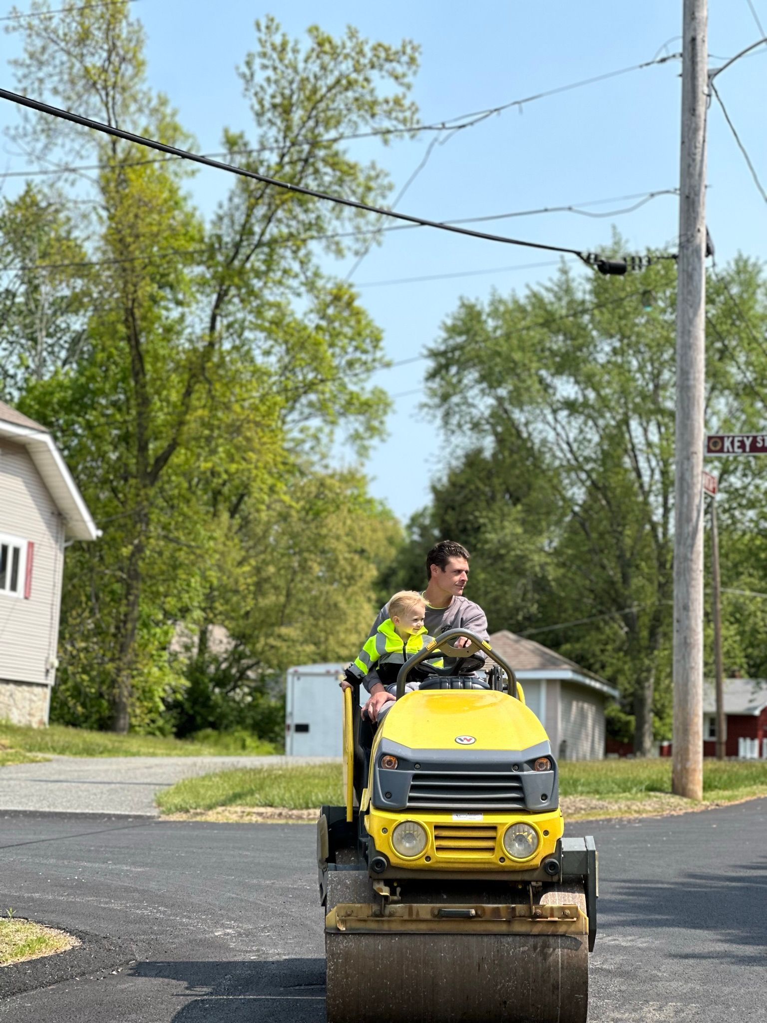 Man and child operating a yellow road roller, outdoors on a paved surface.