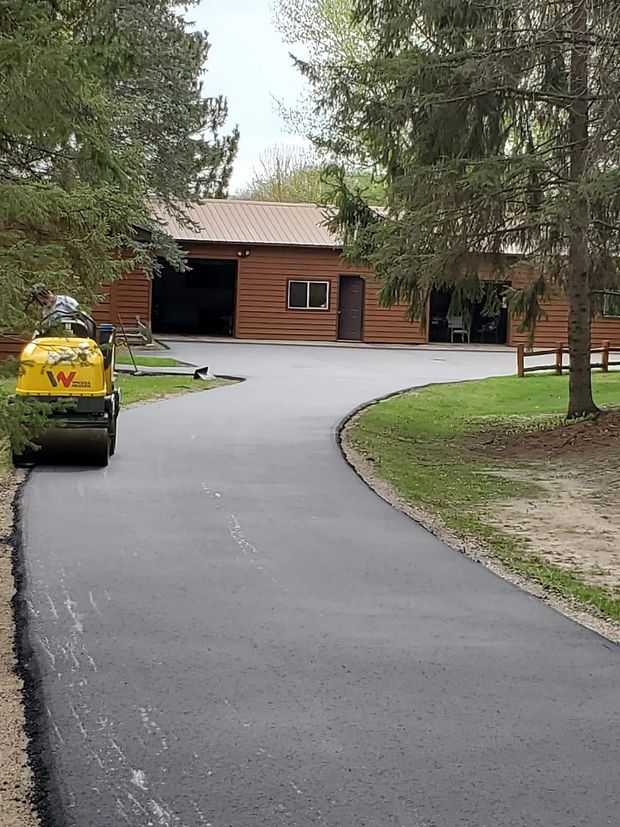 Asphalt driveway leading to a brown building with a roller in the foreground, trees on either side.
