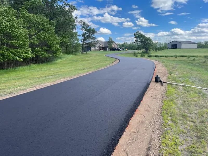 Newly paved asphalt road curves through green landscape, blue sky with clouds.