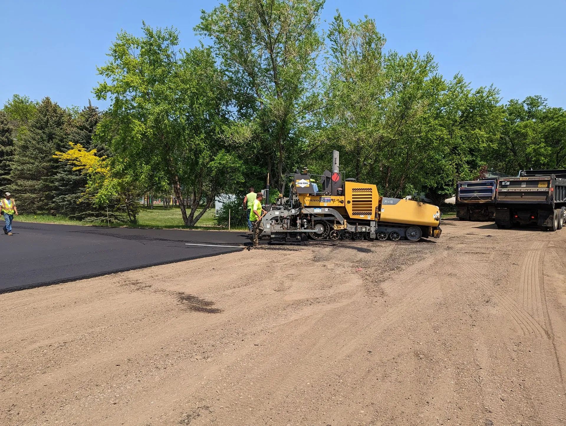 Asphalt paving in progress: machine laying blacktop, workers, dump truck, trees in background.
