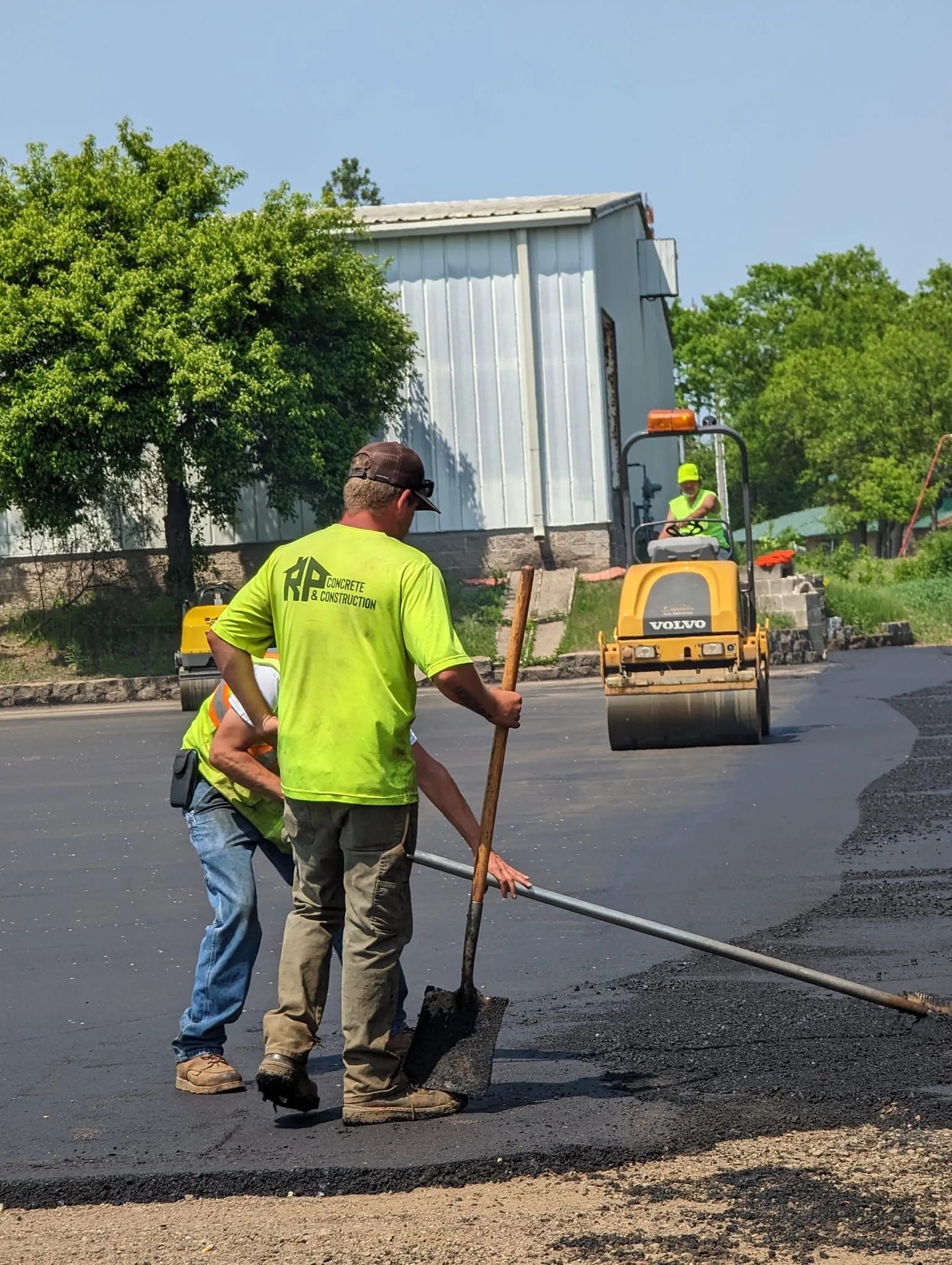 Construction workers paving a road with asphalt. Workers in bright green shirts and one operating a roller.