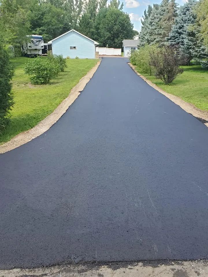 Newly paved, long, dark asphalt driveway leading to light blue garage and houses. Green grass and trees line the sides.