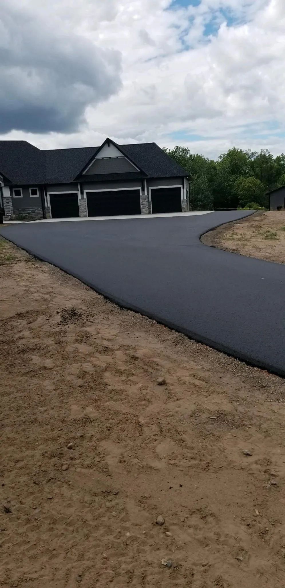 Newly paved black asphalt driveway leading to a house with three garage doors. Brown dirt surrounds the driveway.
