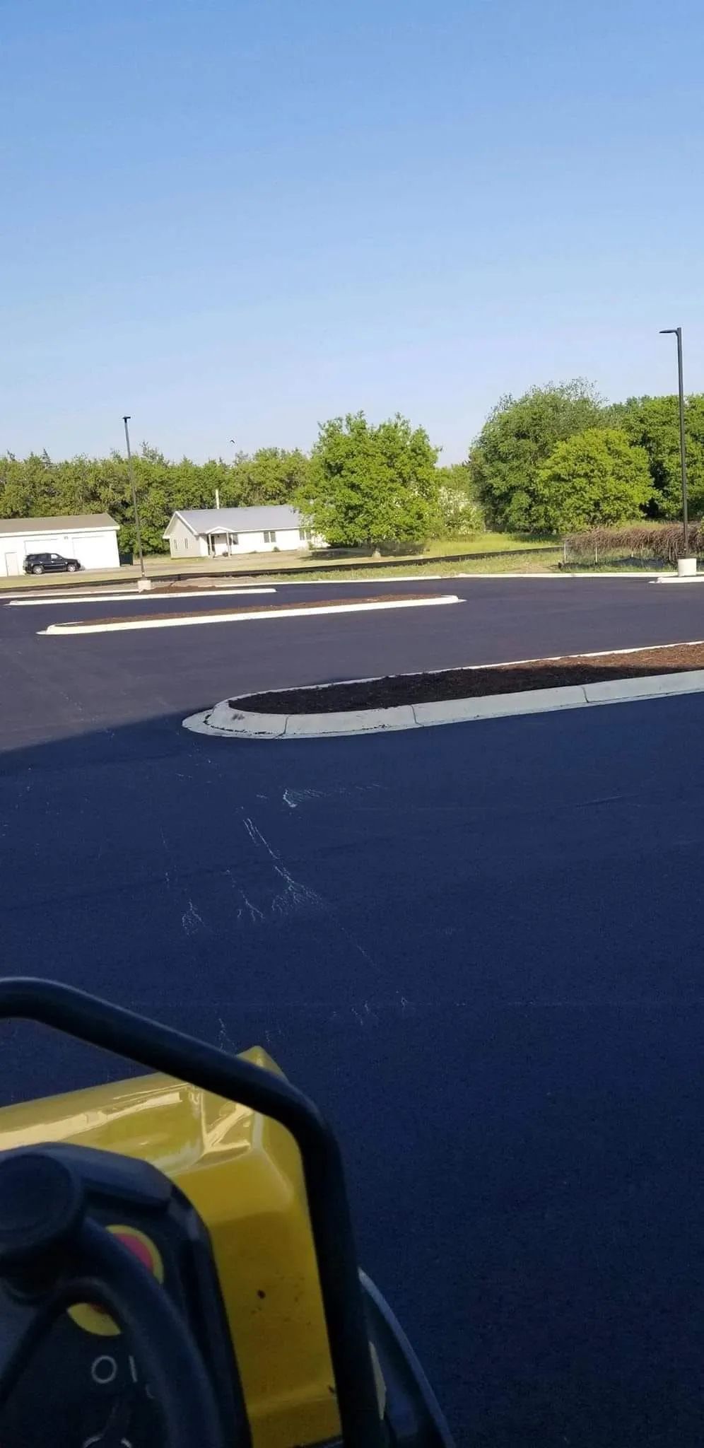 A newly paved parking lot with a yellow compactor in the foreground, trees and a building in the background.